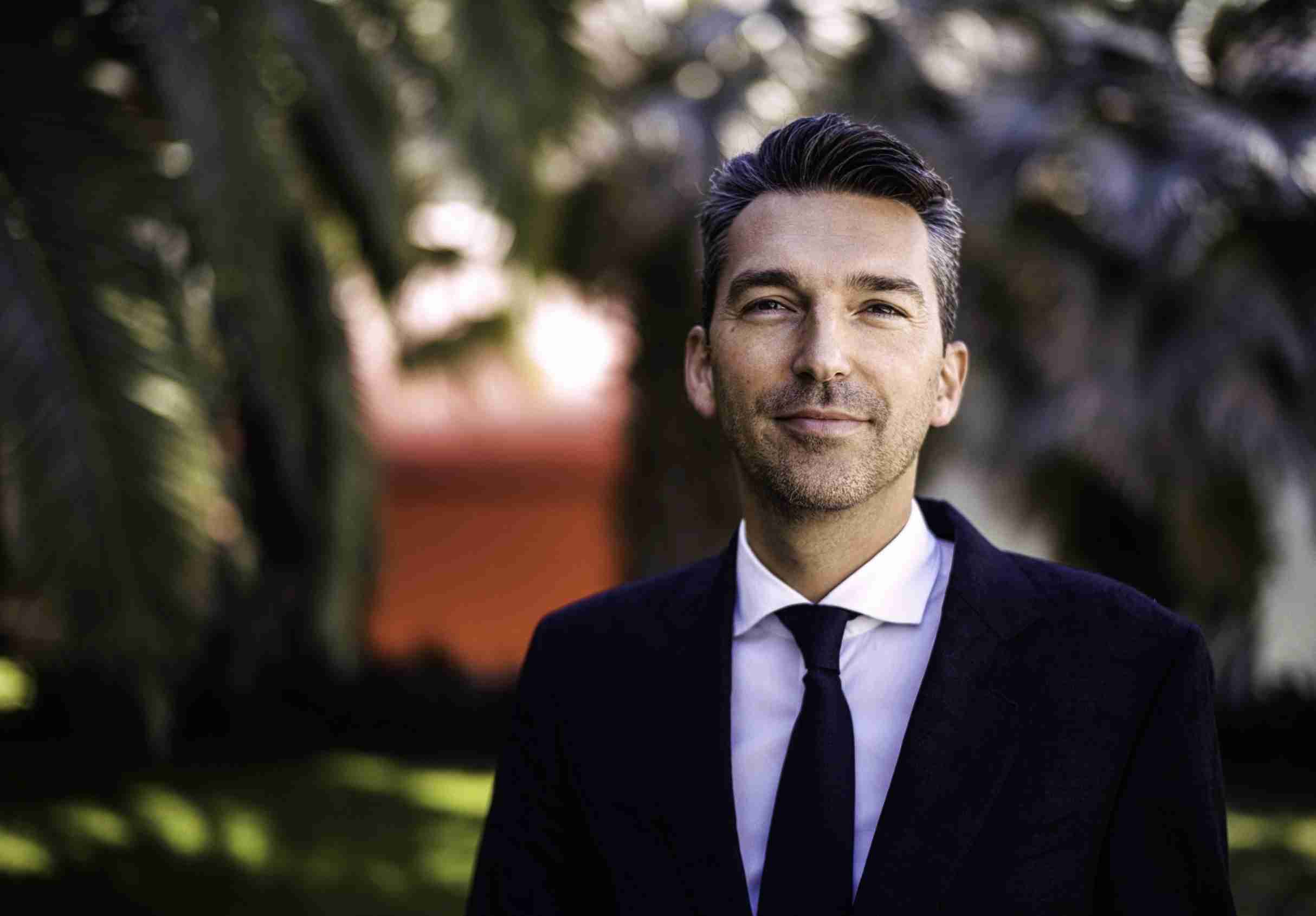 A smiling man is standing confidently in front of a luxury car parked on a tree-lined street.