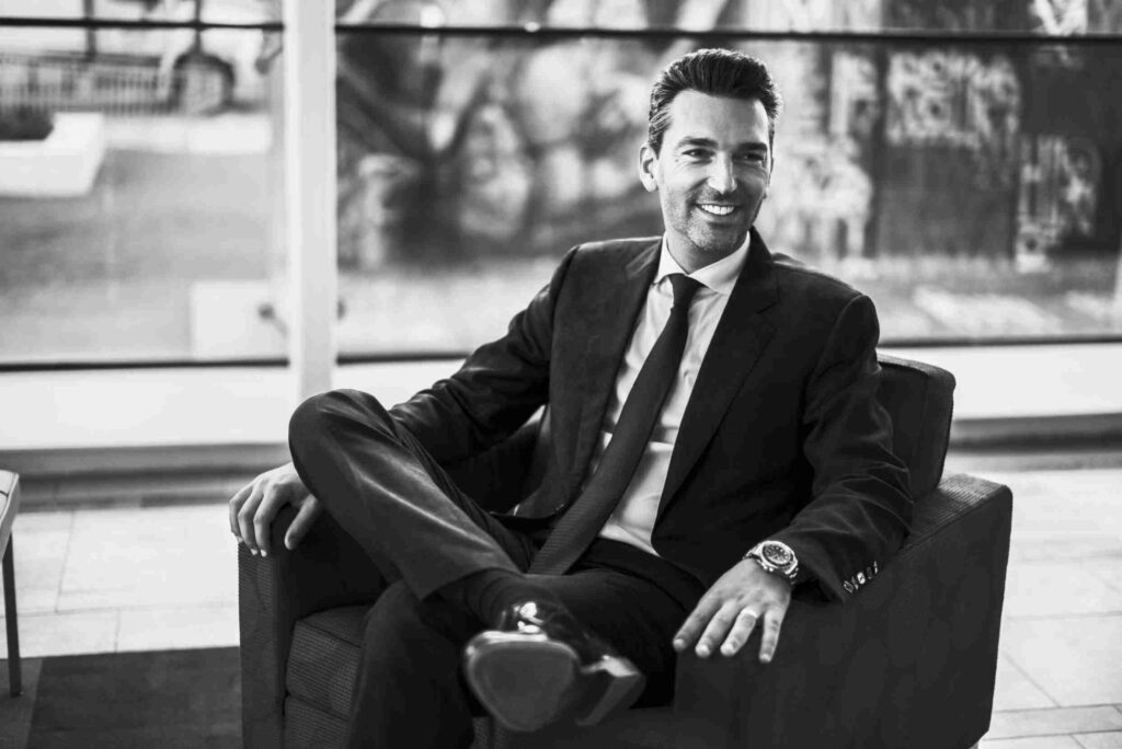 A smiling man is standing confidently in front of a luxury car parked on a tree-lined street.