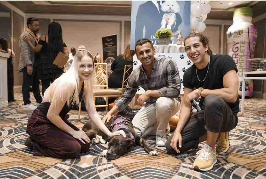 Three people are smiling and posing with a dog at an indoor event.