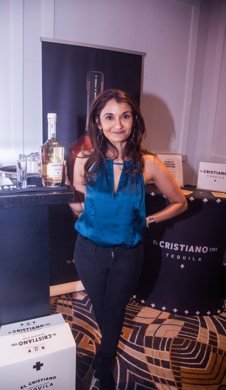 A woman is standing in front of a promotional display for El Cristiano Tequila, smiling at the camera.