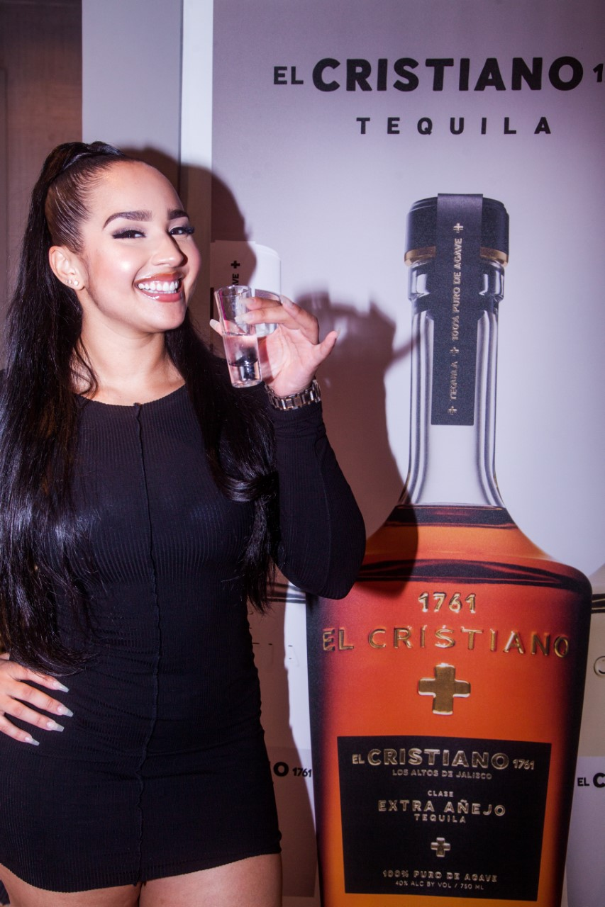 A woman in a black dress is smiling and holding a glass next to a promotional stand for El Cristiano Tequila.