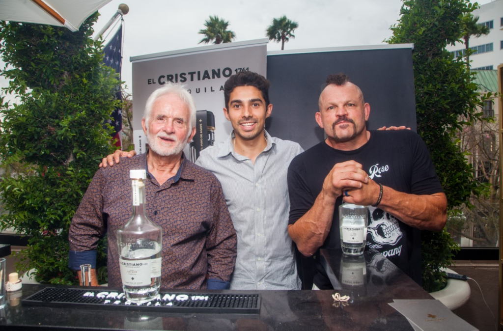 Three smiling men stand behind a bar with bottles of El Cristiano Tequila on display.