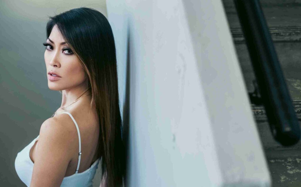 A man in glasses and a blue suit poses with his hands up against a backdrop with text overlays referring to entertainment.