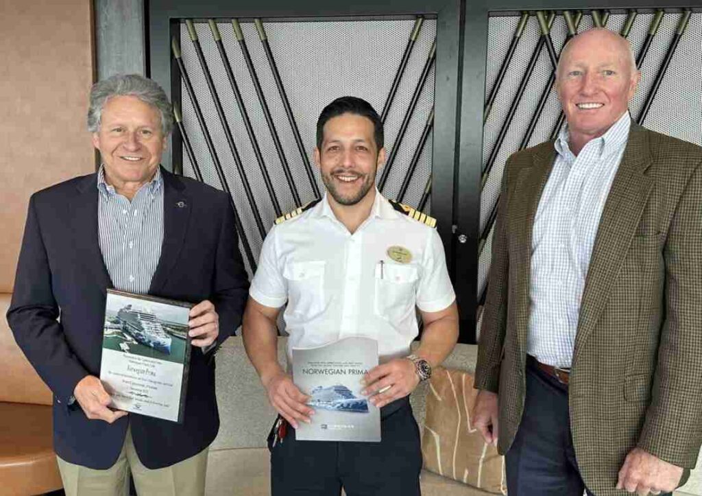 Three men are smiling for a photo; the one in the middle is in a white naval uniform, holding a framed certificate, flanked by two in business attire.