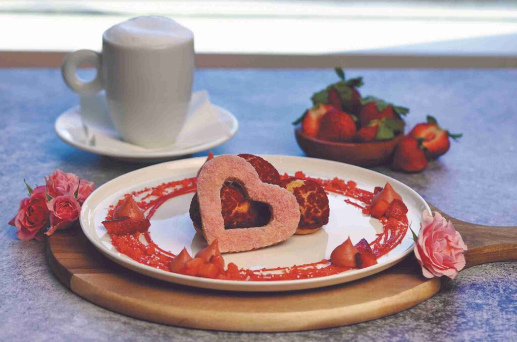 A romantic breakfast setup featuring a heart-shaped egg, pastries, strawberries, flowers, and a cup of cappuccino on a wooden tray.