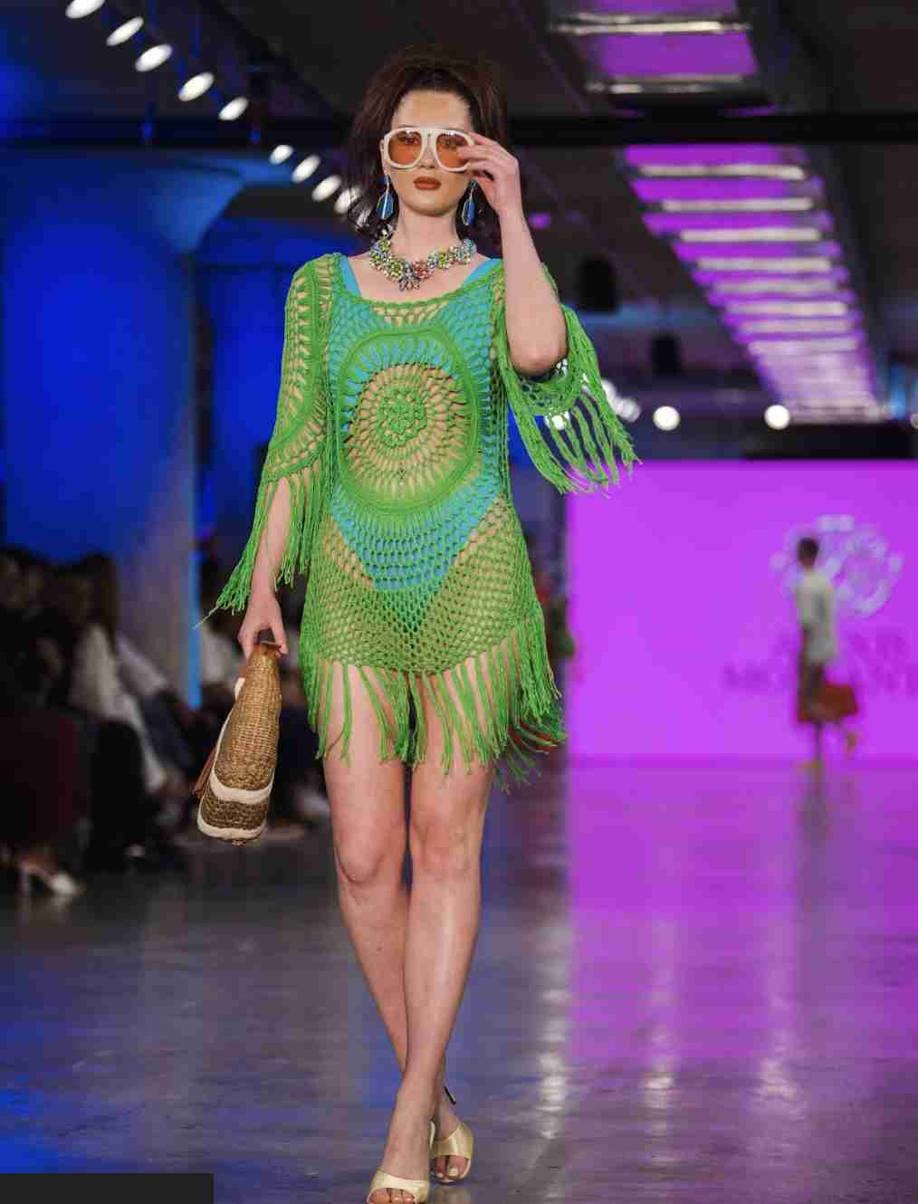 A model is walking down the runway showcasing a vibrant green knit dress with fringe details, accessorized with sunglasses, a necklace, and a handbag.