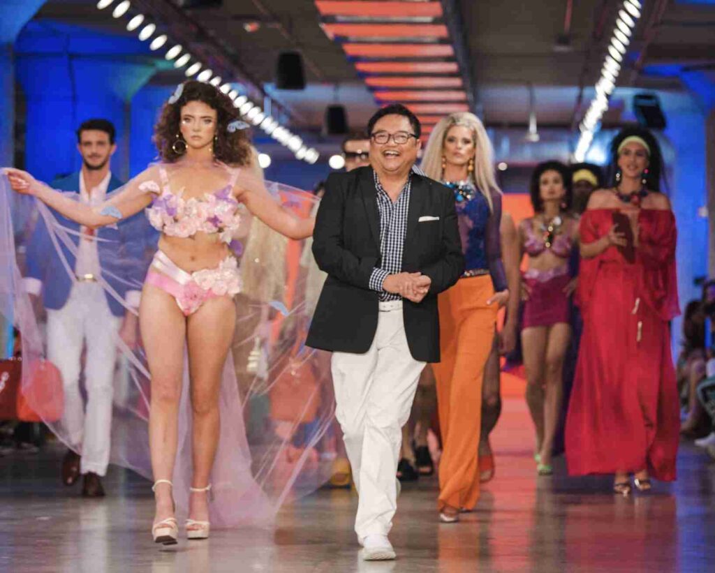 A model is walking down the runway showcasing a vibrant green knit dress with fringe details, accessorized with sunglasses, a necklace, and a handbag.