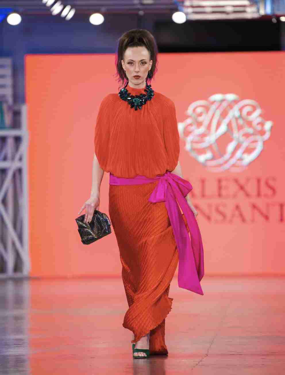 A model is walking down the runway showcasing a vibrant green knit dress with fringe details, accessorized with sunglasses, a necklace, and a handbag.