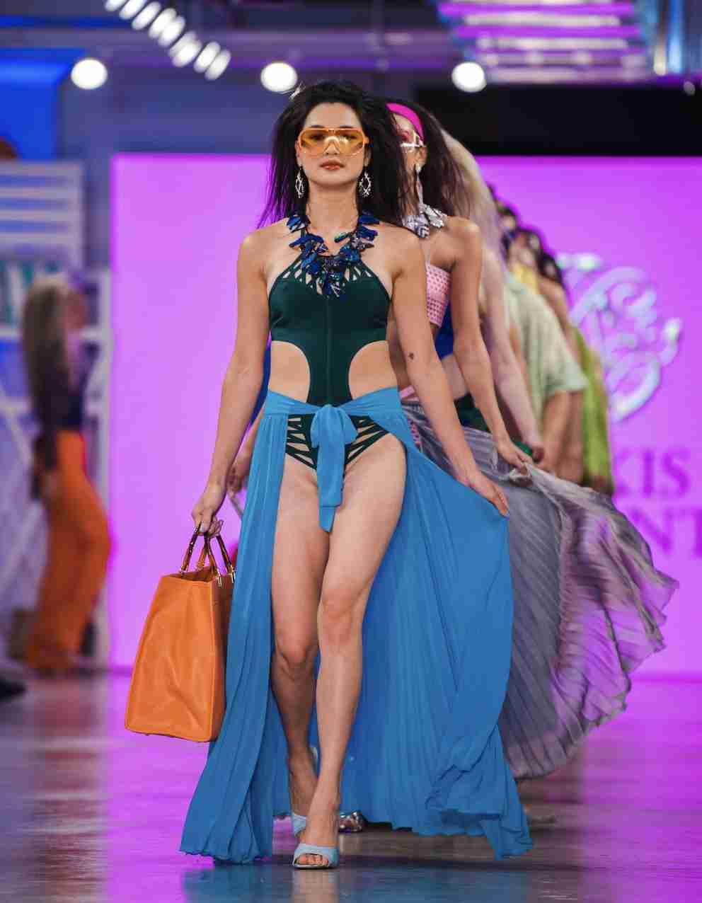 A model is walking down the runway showcasing a vibrant green knit dress with fringe details, accessorized with sunglasses, a necklace, and a handbag.
