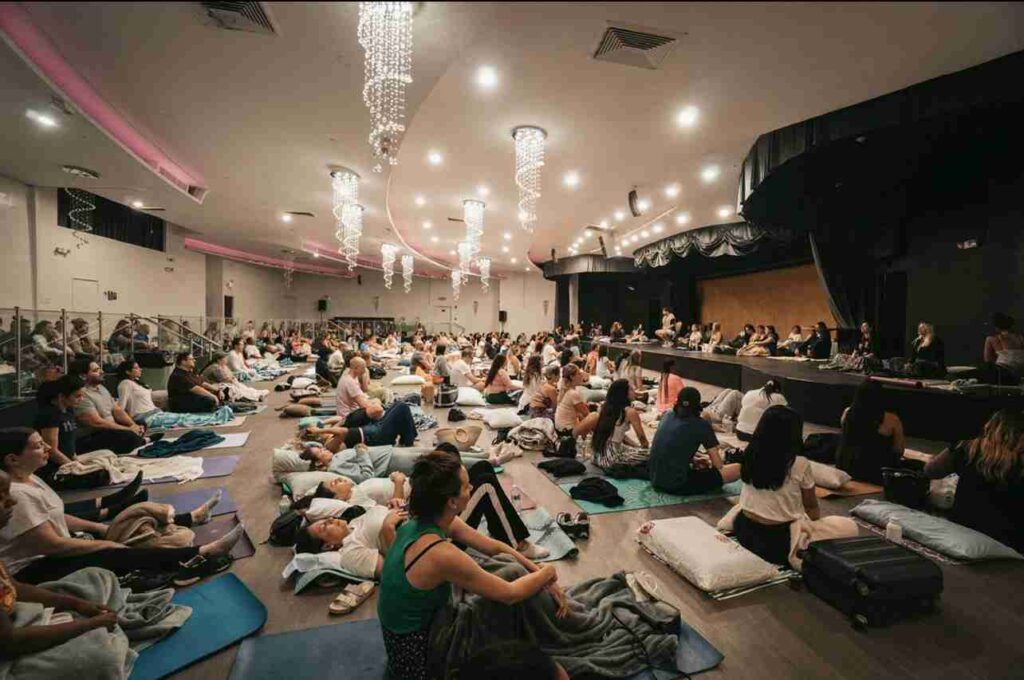 A group of people sitting on mats and pillows attentively watching a presentation in a large, modern hall with decorative lighting.