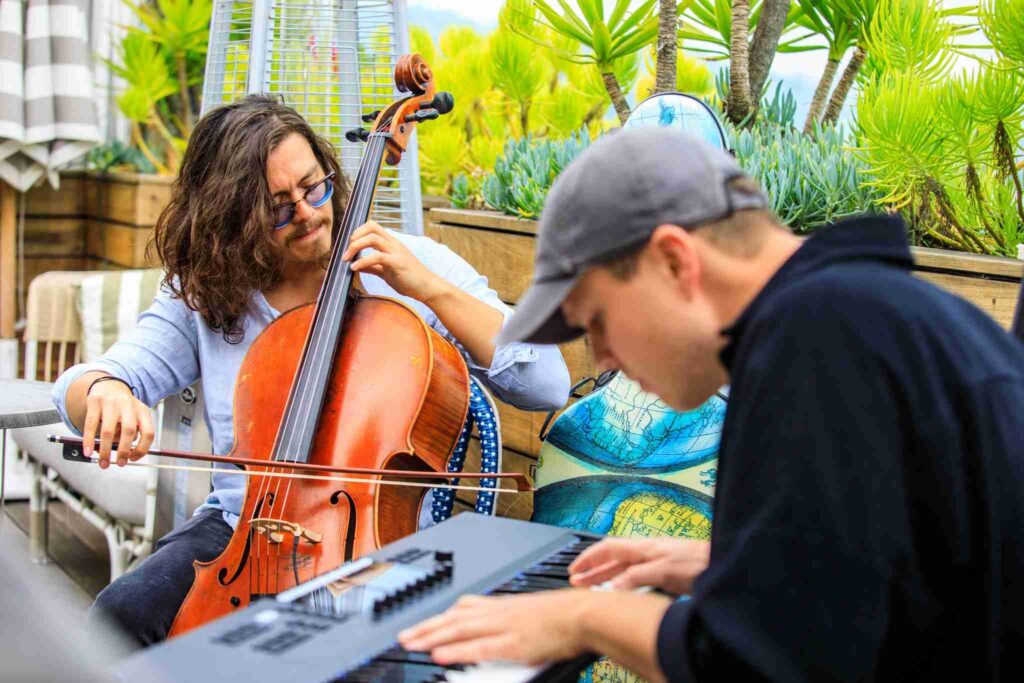 Two musicians are playing instruments, a cello and a keyboard, in an outdoor setting with greenery around.