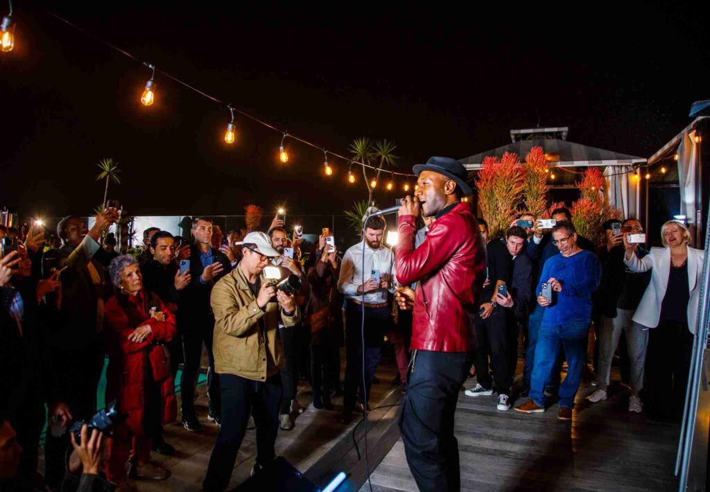 A man performs enthusiastically at an outdoor evening event while surrounded by an audience capturing the moment with their phones.