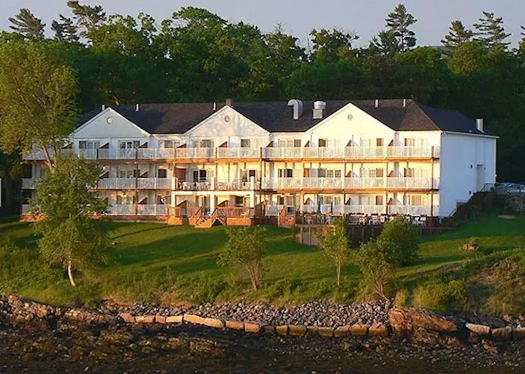 A large waterfront hotel with multiple balconies bathed in warm sunlight, surrounded by greenery and a rocky shoreline.