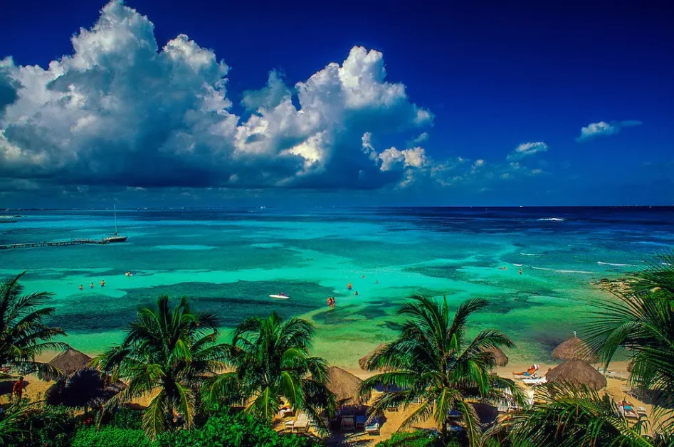 A tropical beach with clear blue water, palm trees, and people enjoying various water activities under a bright sky with fluffy clouds.