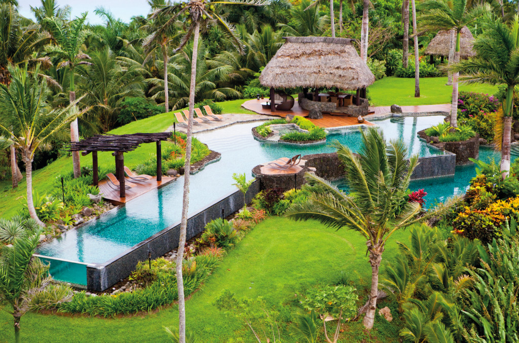 A tropical resort with a thatched-roof hut, an infinity pool surrounded by palm trees, and lush green landscaping.