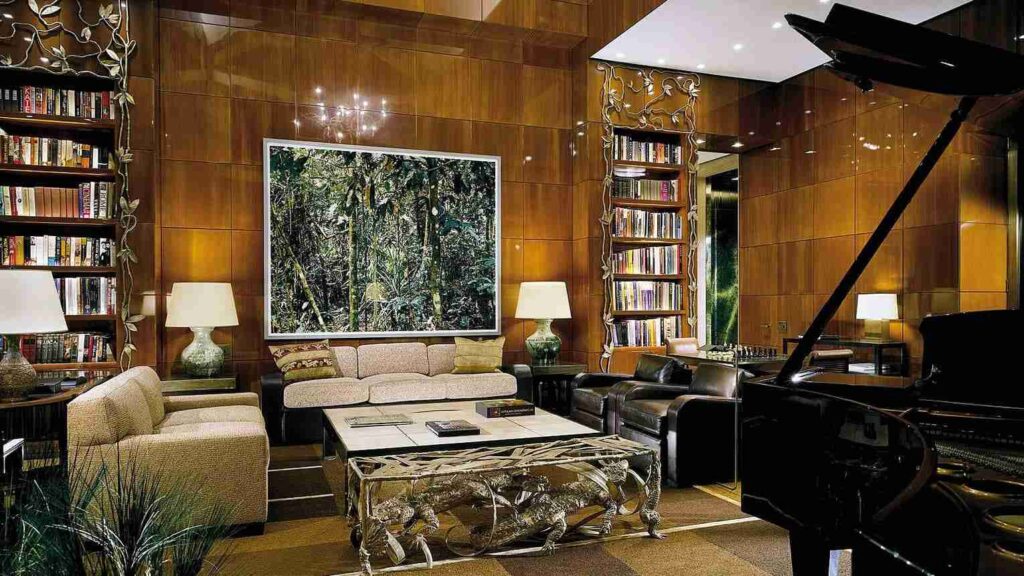 An elegant living room with a grand piano, plush seating, bookshelves, and a large painting on the wood-paneled wall.