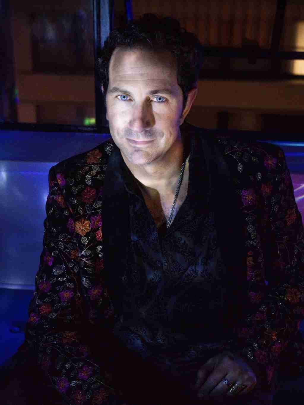 A man in a light green suit stands with his hands over his chest against a grey background.