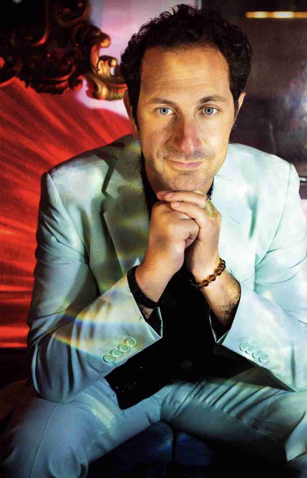 A man in a light green suit stands with his hands over his chest against a grey background.