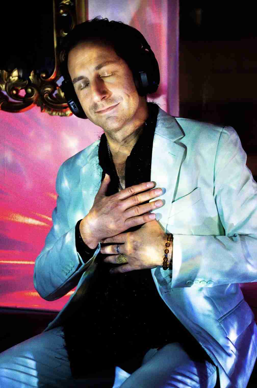 A man in a light green suit stands with his hands over his chest against a grey background.