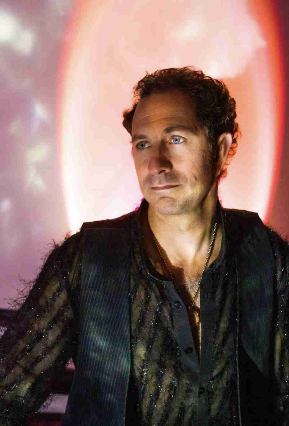 A man in a light green suit stands with his hands over his chest against a grey background.