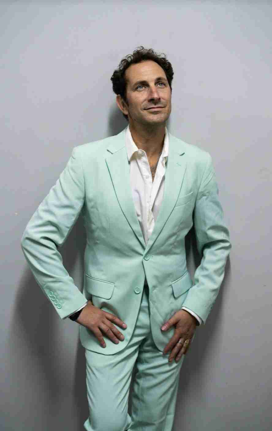 A man in a light green suit stands with his hands over his chest against a grey background.