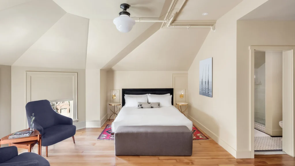 A well-lit, cozy attic bedroom with a sloped ceiling, featuring a queen bed, seating area, decorative rug, and an adjoining bathroom.