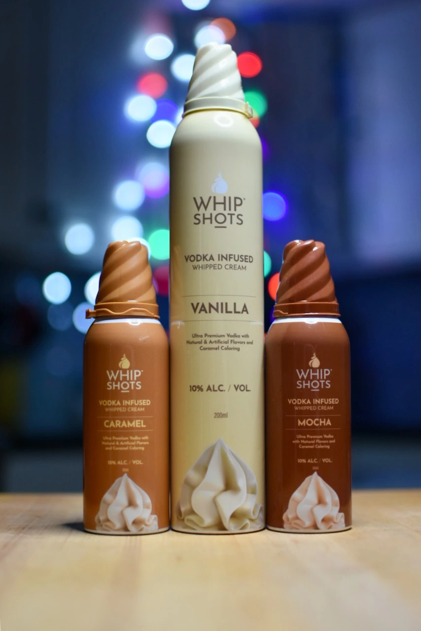 Three bottles of alcohol-infused whipped cream with varying flavors, prominently displayed with colorful blurred lights in the background.