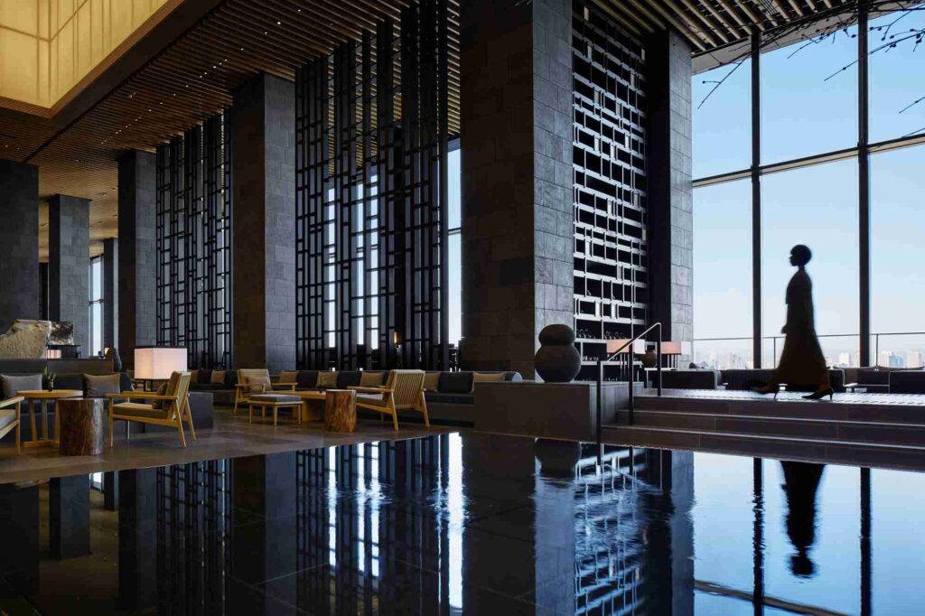 A person walking through a spacious, modern hotel lobby with high ceilings, large windows, and reflective floors.