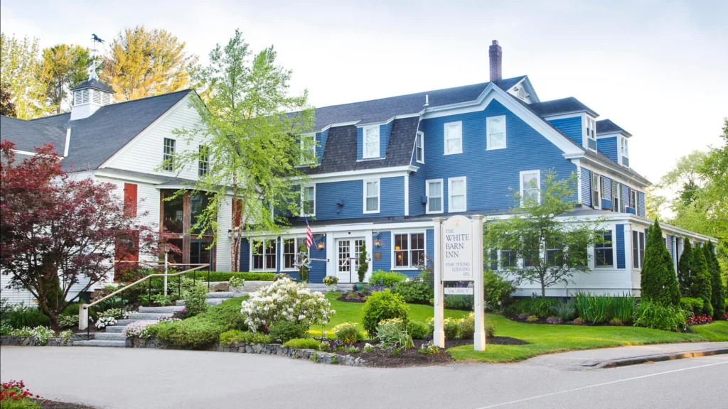 A large blue two-story inn with a signpost in front surrounded by well-manicured greenery and a clear sky.