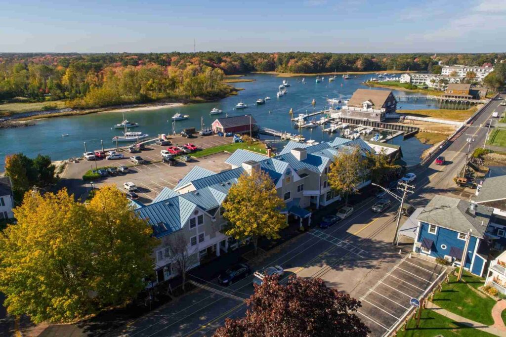 An aerial view of a coastal town featuring buildings, a marina with boats, and a river lined with autumn trees.