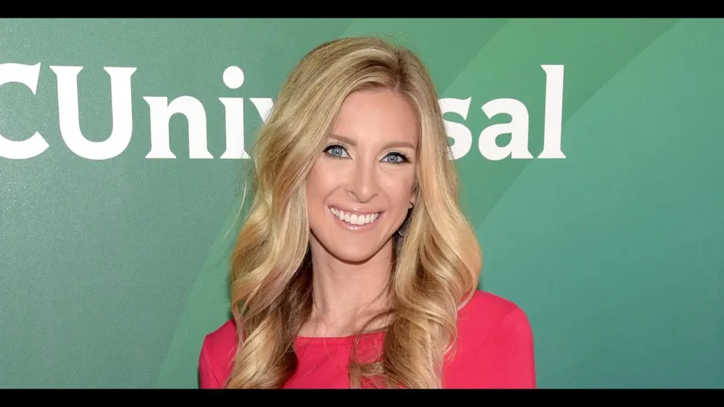 A smiling woman with blonde hair wearing a red dress at an event with a green and white NBCUniversal backdrop.