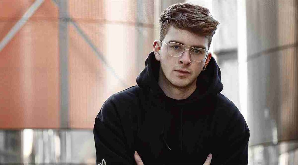 A young man with glasses wearing a black hoodie is posing with his arms crossed against an urban backdrop with orange elements.