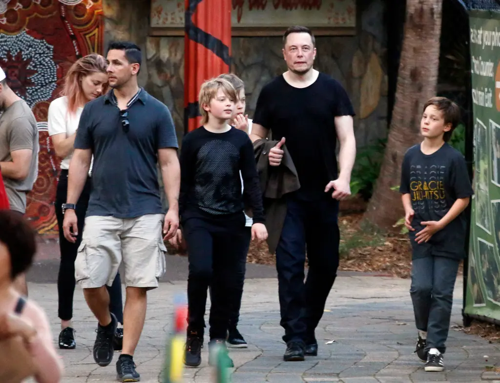 The image shows a group of people casually walking on a path in an outdoor setting, with trees in the background.