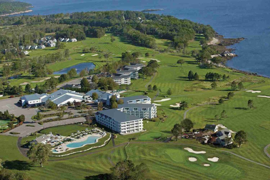 An aerial view of a coastal resort with a golf course, buildings, and pools surrounded by greenery and blue waters.