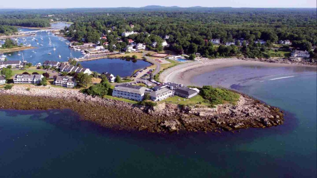 An aerial view of a coastal area showing buildings, a curved beach, trees, and boats near a calm blue bay.