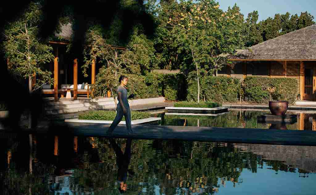 A person is walking by a reflective pond in a serene garden with traditional-style buildings in the background.