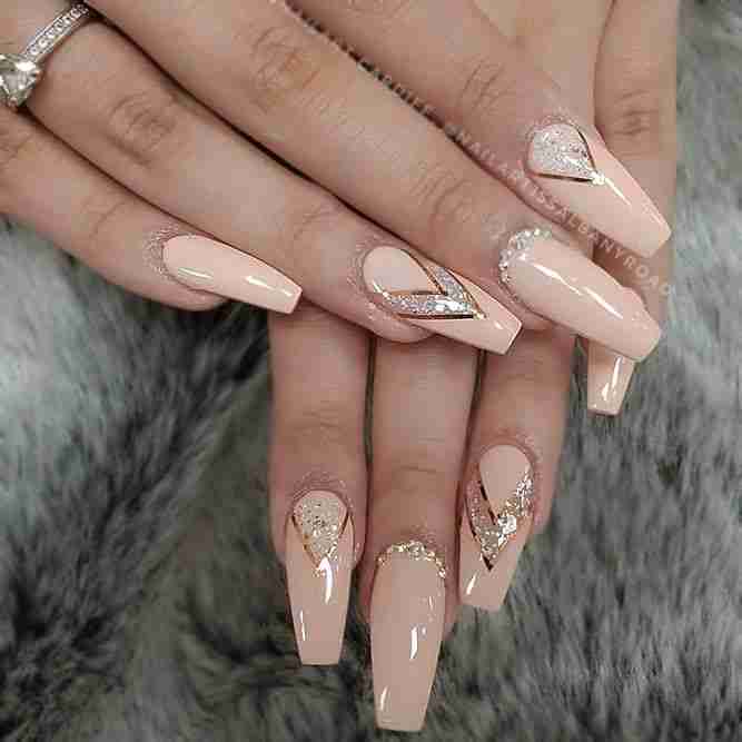 A set of hands showcasing long, nude-colored nails with glitter and jewel accents, against a furry gray background.