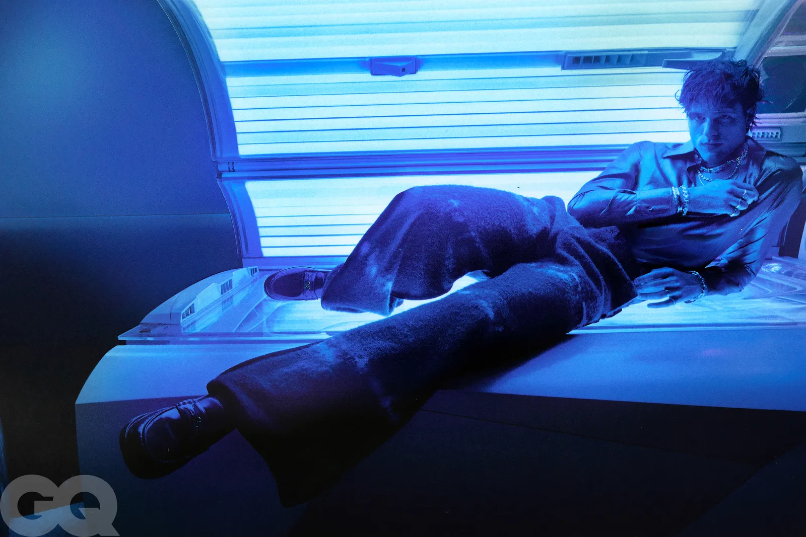 A man lounges atop a tanning bed under blue light, dressed in a suit with a relaxed pose for a stylized photoshoot.