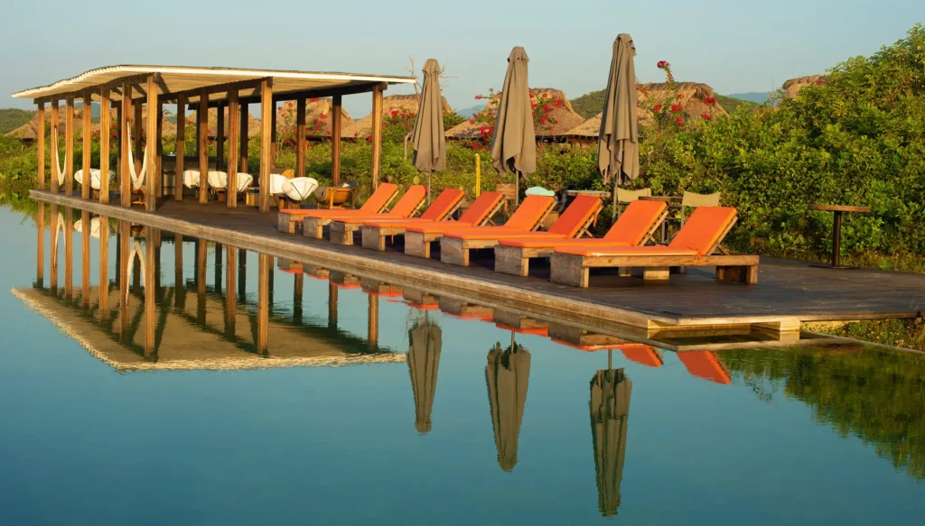 A serene outdoor poolside setting with lounge chairs, umbrellas, and a wooden deck overlooking a calm body of water.