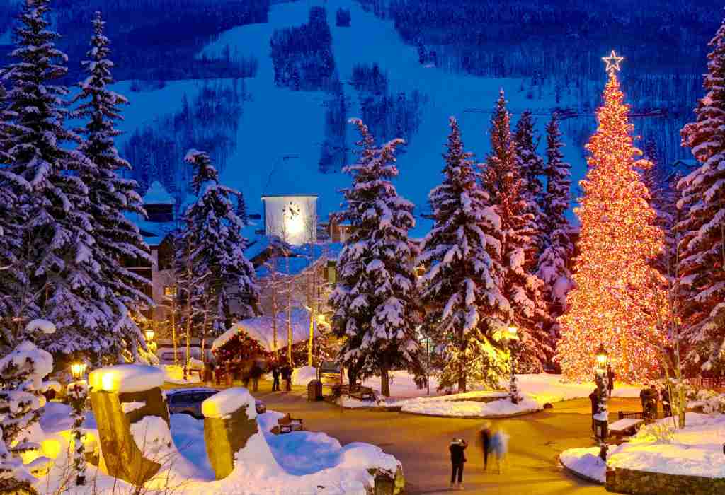 A festive winter scene with snow-covered trees, a brightly lit Christmas tree, and people walking in a picturesque mountain resort town at dusk.