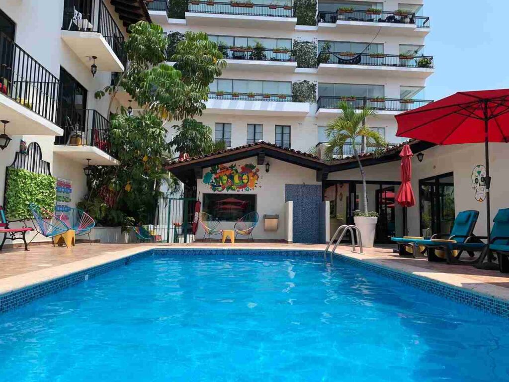 A vibrant poolside area with loungers and a red umbrella, fronting a building with balconies and a colorful, artistic sign.