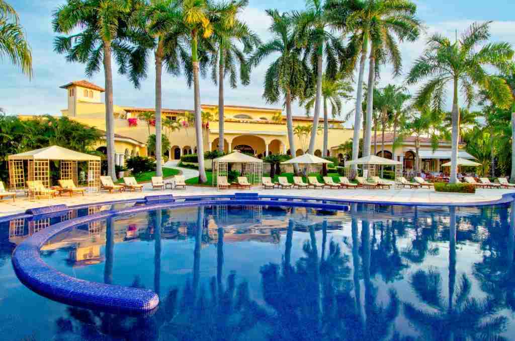 A luxurious resort with a large swimming pool surrounded by palm trees under a clear sky at dusk.