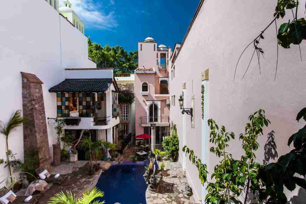 The image displays a charming boutique hotel courtyard with a small pool, surrounded by multi-story buildings with vibrant architectural details and lush greenery.
