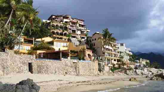 A coastal scene with a sandy beach in the foreground and multicolored buildings on a hillside under a cloudy sky.