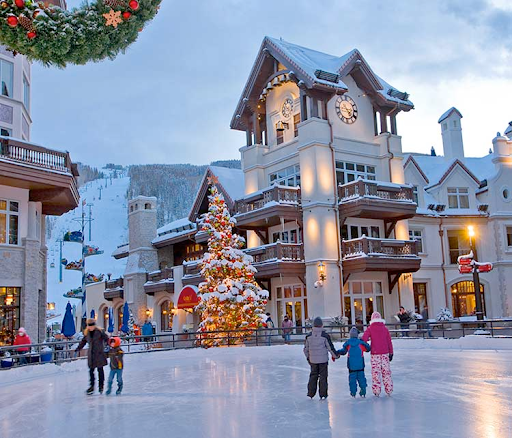 An outdoor ice skating rink with people skating in front of a festively decorated building with a Christmas tree during evening.