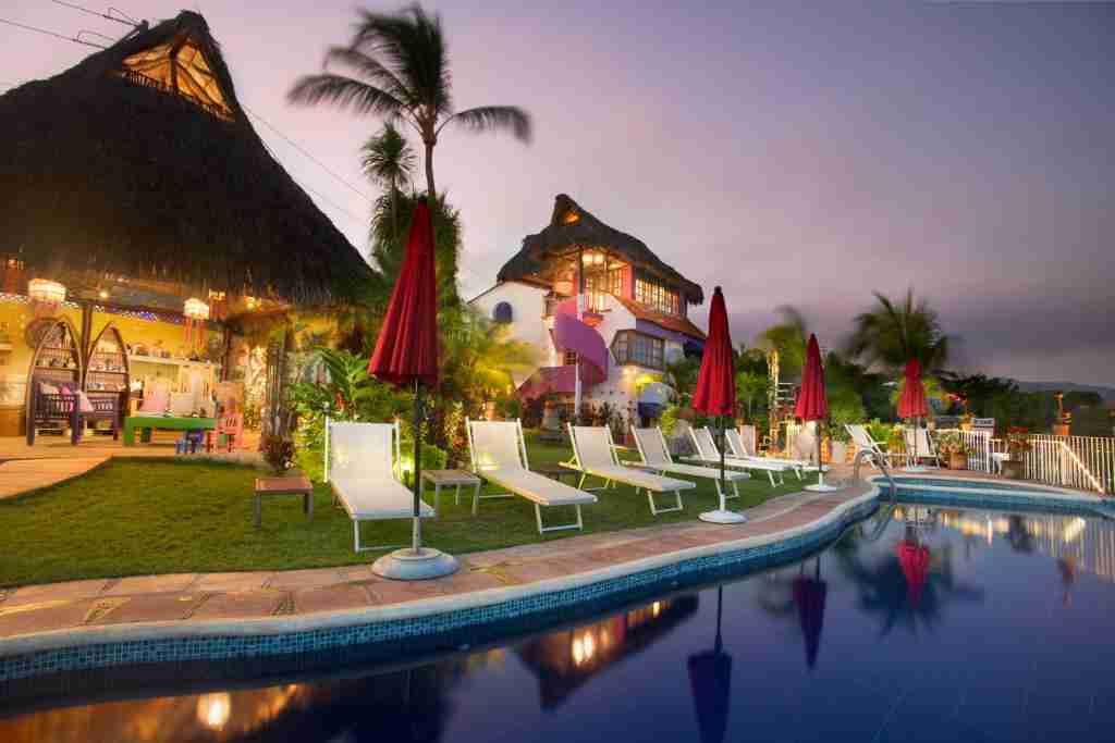 A tranquil evening scene at a resort with lounge chairs by a pool, a thatched-roof structure, and a warmly lit building under a dusk sky.