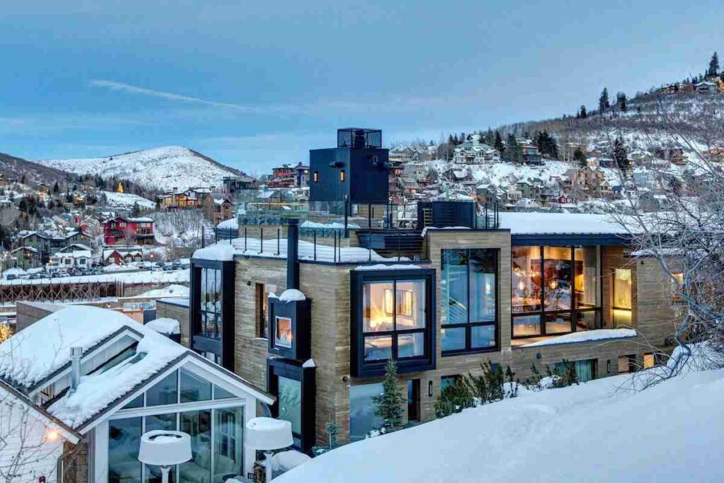 A modern house with lit interior on a snowy hillside with a view of a wintry landscape and other houses in the distance.
