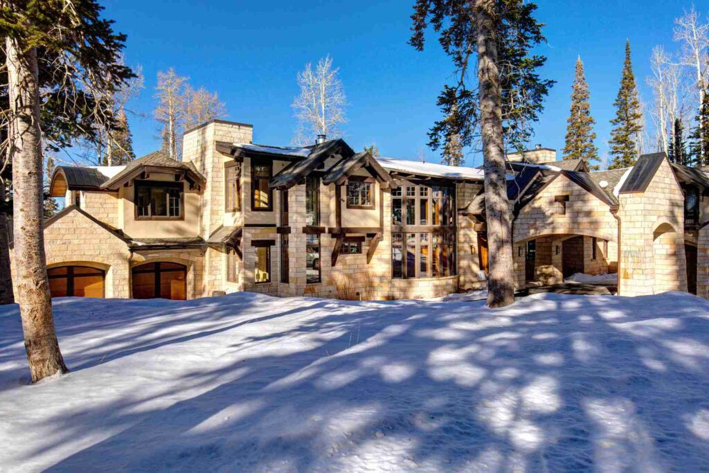 A luxurious stone house with large windows, surrounded by snow-covered trees under a clear blue sky.