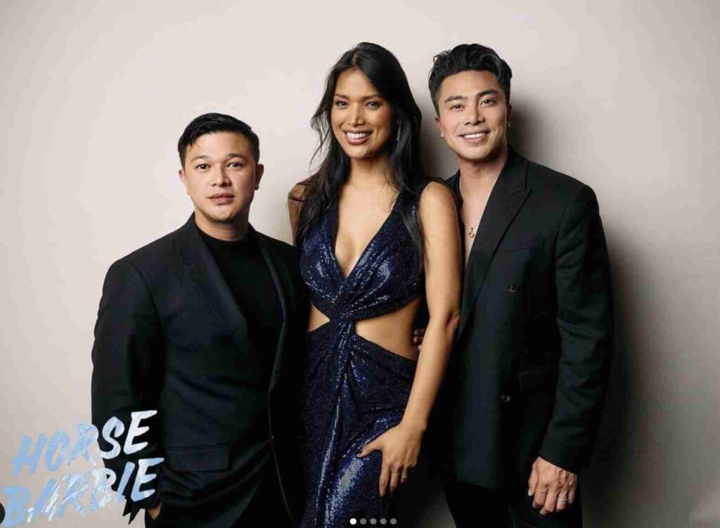 A man poses on the red carpet wearing a stylish red patterned blazer over a black shirt and trousers, paired with black boots.
