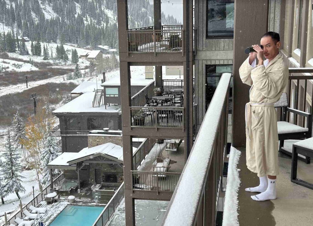 A person is relaxing in a steaming outdoor hot tub with a snowy mountain backdrop under a clear sky.